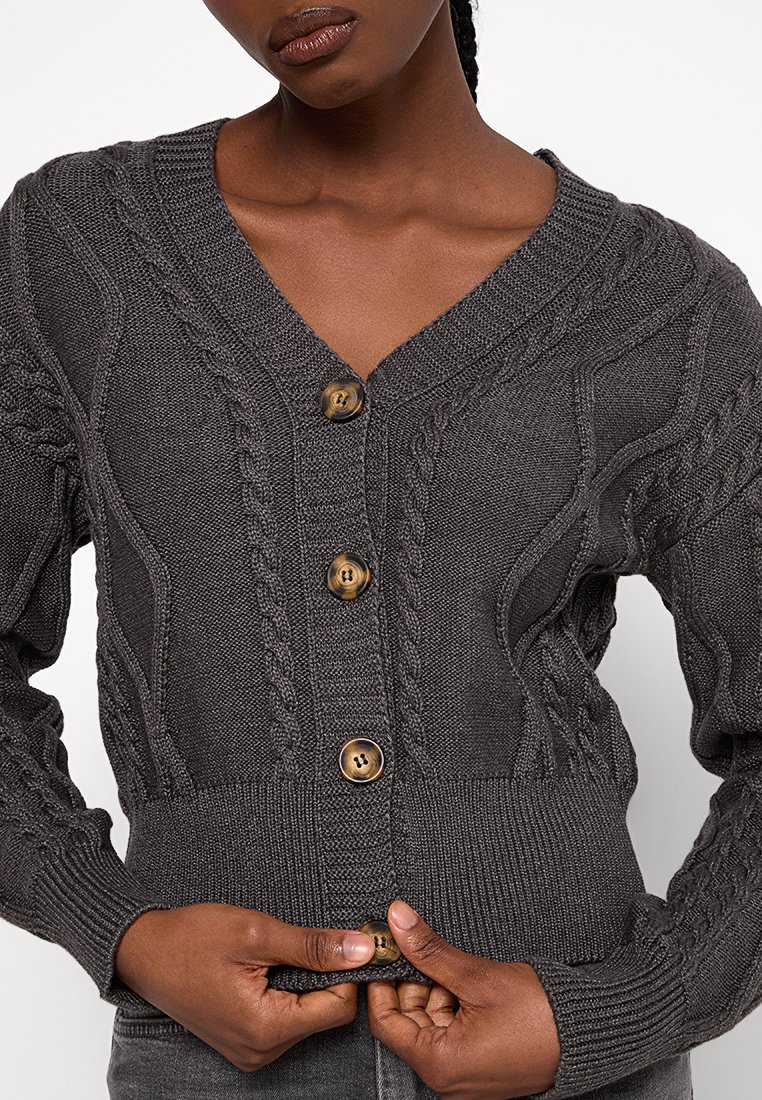 Grijs kabelgebreid vest met een V-hals, zes knopen, geribbelde onderrand en getextureerde mouwen. Voorzien van grote, ronde bruine knopen voor sluiting.