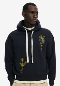 Marineblå hættebluse med hvide snørebånd, prydet med gul blomsterbroderi og en afslappet pasform. Glat stof med ribbede kanter og manchetter.