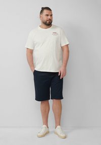 Weißes Baumwoll-T-Shirt mit rundem Ausschnitt und rotem Drucktext, kombiniert mit dunkelblauen knie-langen Shorts und weißen Turnschuhen.