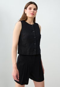 Femme portant un haut noir sans manches avec boutons et un short noir plissé, debout devant un fond gris clair uni.