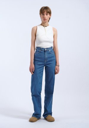 Persona in piedi che indossa una canotta bianca senza maniche, jeans blu a vita alta, scarpe color cuoio e collane a strati su uno sfondo chiaro e semplice.