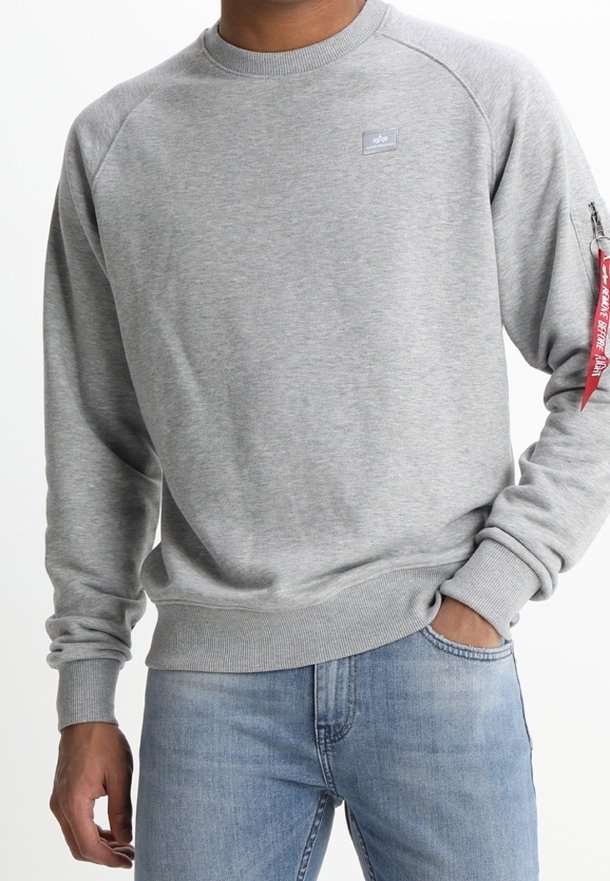 Homme portant un sweat-shirt gris clair avec un petit écusson sur la poitrine et une étiquette rouge sur la fermeture éclair de la manche, associé à un jean bleu clair, main dans la poche.