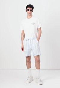 T-shirt blanche à manches courtes avec un logo imprimé, associée à un short rayé bleu clair et blanc. Le mannequin porte des lunettes de soleil et des baskets blanches.