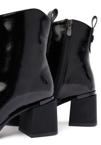 Botines negros hasta el tobillo con un acabado brillante de charol, tacones cuadrados de bloque y cremallera lateral, que presentan una textura suave y mínimos acentos de hardware.