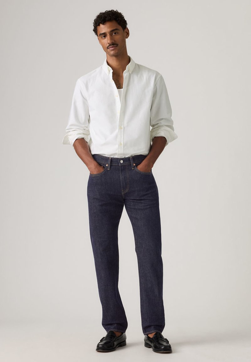 Hombre de cabello rizado vistiendo una camisa blanca abotonada, jeans azul oscuro y mocasines negros, de pie con las manos en los bolsillos.