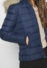 Kvinna som bär en marinblå quiltad jacka med pälskantad huva, en beige stickad tröja, ett guldhalsband och svarta byxor med handen i fickan.