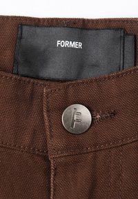 Brune bomuldsbukser med en sølvmetalknap med et præget "F"-logo og et sort stofmærke, der viser mærkenavnet "FORMER."