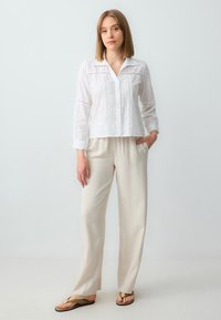 Witte, knooploze blouse met oogjesdetails en lange mouwen, gecombineerd met lichtbeige, wijde broekspijpen. Model staat met één hand in de zak.