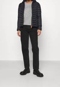 Person iført en sort pufferjakke over en grå sweater, sorte lige ben jeans og sorte ankelstøvler, stående foran en ensfarvet baggrund.