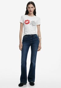 T-shirt en coton blanc avec un graphisme de lèvres rouges et du texte noir, associé à un jean évasé bleu foncé et des chaussures noires à enfiler.