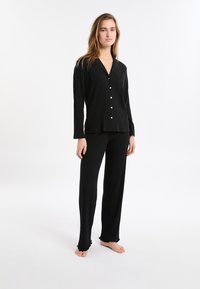 Czarny komplet piżamy z żebrowanym wzorem, składający się z koszuli zapinanej na guziki z kieszonką oraz długich spodni z falbanami na końcach. Wykonany z miękkiego materiału dla zapewnienia komfortu.