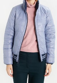 Světle modrá prošívaná bunda se zipovým zapínáním, žebrovanými námořnickými ozdobami na lemu a manžetách a stojatým límcem přes růžový svetr.