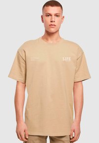 Beige bomulds t-shirt med løs pasform, korte ærmer, rund halsudskæring og fronttryk i hvid. Teksten inkluderer ordet "LIFE."