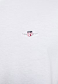 Witte stof met een klein geborduurd logo met een schild met rode en witte strepen en drie witte sterren op een blauwe achtergrond.