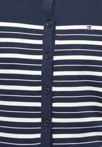 Ναυτικό μπλε ζακέτα με οριζόντιες λευκές ρίγες, με στρογγυρούς, σκούρους κουμπιά και μια μικρή λογότυπη λεπτομέρεια στην αριστερή πλευρά. Μαλακή πλεκτή υφή.