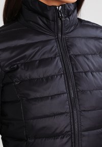 Sort polstret jakke med høj krave, der har en quiltet tekstur og en frontlynlås. Glat materiale med en subtil glans.