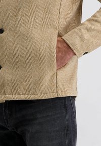 De hand van een man in de zak van een beige jasje met visgraatpatroon, gekleed in zwarte jeans, tegen een effen lichte achtergrond.