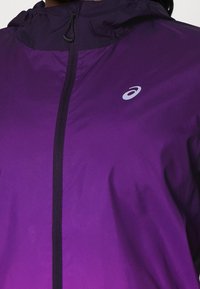 Chaqueta ligera morada con capucha, con cierre frontal y logo reflectante. Textura suave y diseño minimalista.