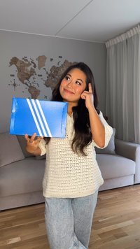 Zils kartona kastīte ar baltu trīs svītru dizainu, ko tur cilvēks krēmkrāsas adītā džemperī, stāvot telpā ar koka grīdu un kartes sienu.