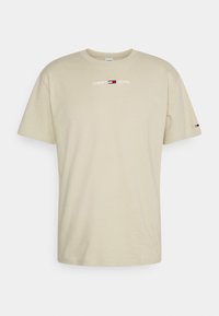 Světle béžové bavlněné tričko s krátkým rukávem, s potiskem loga "TOMMY JEANS" v bílé barvě a červeno-modrou nášivkou na levém rukávu.