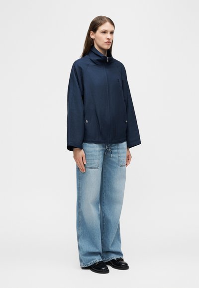 Γυναίκα στέκεται με το πρόσωπο προς τα εμπρός, φορώντας μπλε σακάκι με φερμουάρ, φαρδιά ανοιχτό μπλε τζιν με μεγάλες μπροστινές τσέπες και μαύρα παπούτσια σε απλό φόντο.