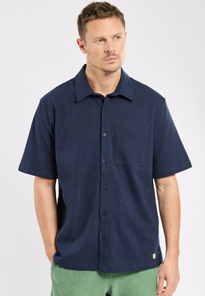 Homme aux cheveux courts portant une chemise à manches courtes boutonnée texturée bleu foncé et un pantalon vert clair, regardant sur le côté.