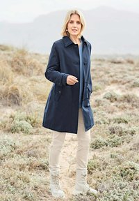Navy trench coat with button closure, side pockets, and a smooth texture. Paired with light beige pants and lace-up beige boots.