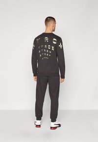Schwarzer Hoodie mit buntem "HYROX"-Text und Designdetails, kombiniert mit passenden Jogginghosen. Modell trägt weiße und rote Sneakers.