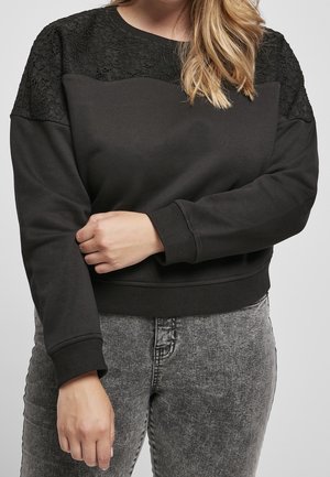Femme portant un sweat-shirt noir à manches longues avec un détail en dentelle aux épaules, associé à un jean gris délavé à l'acide, tenant un bras croisé sur son corps.