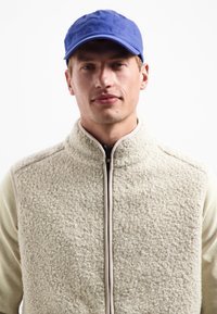 Man in blauwe pet en beige textuur zip-up fleece vest over een lichtkleurige lange mouw shirt, neutrale uitdrukking, witte achtergrond.