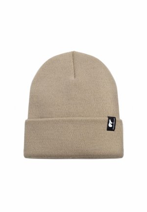 Tans kleurige gebreide beanie met een omgeslagen rand, voorzien van een klein zwart label aan de zijkant. Het materiaal lijkt zacht en rekbaar.