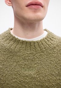 Maglione a maglia verde oliva con una finitura testurizzata e pelosa, dal collo a giro. Presenta un logo "Hugo" in rilievo sulla parte frontale.