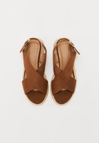 Brown suede sandals with crisscross straps, a flat espadrille sole, and a metal buckle detail on the side. Soft texture and open toe design.