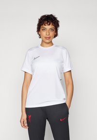 Biela atletická športová tričko s krátkym rukávom, okrúhlym výstrihom a čiernym logom Nike na hrudi, kombinované s tmavošedými nohavicami s červeným logom.