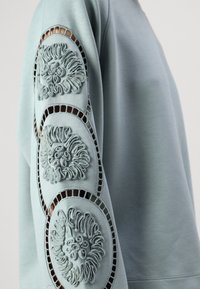 Sweat-shirt bleu clair présentant trois motifs de lion en relief sur les manches, avec des détails découpés et une texture lisse.