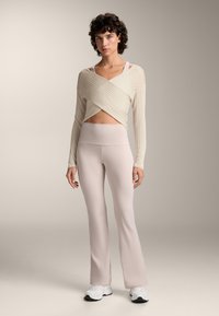 Hellbeige Wickeloberteil mit gerippter Textur, kombiniert mit blassen, pinken Schlagleggings. Das Model trägt weiße Sportschuhe und steht vor einem neutralen Hintergrund.