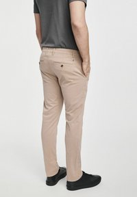 Beżowe chinosy o slim fit, z dwiema tylnymi kieszeniami z zapięciem na guziki, noszone z ciemnym t-shirtem i czarnymi trampkami.