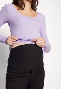 Top de manga larga de color morado claro con cuello en forma de scoop y detalle de botones, combinado con pantalones de maternidad negros y elásticos.