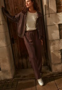 Bruine bomberjack met ritsdetails, beige shirt en bijpassende bruine sweatpants. Zilveren sneakers maken de outfit compleet. Achtergrond van hout en steen.