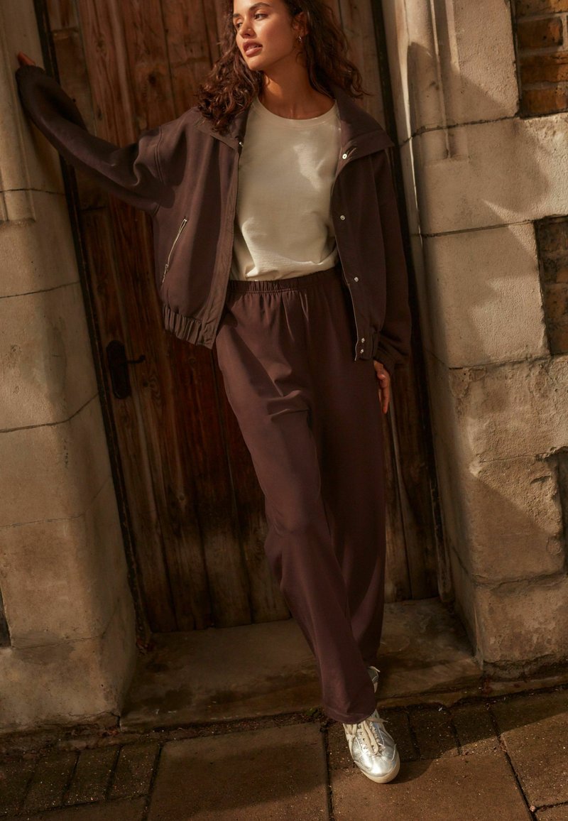 Bruine bomberjack met ritsdetails, beige shirt en bijpassende bruine sweatpants. Zilveren sneakers maken de outfit compleet. Achtergrond van hout en steen.