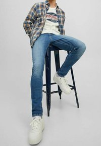 Jeans de mezclilla azul, zapatillas blancas y una camisa de cuadros en marrón y azul sobre una camiseta gráfica vintage blanca. Sentado en un taburete negro.