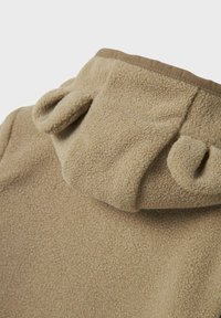 Beige fleece tyger med huva som har två små, rundade djurliknande öron sydda upptill.
