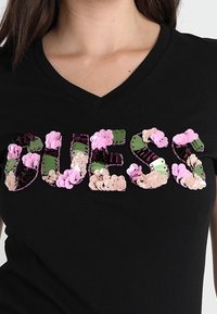Musta V-pääntie T-paita, jossa on "GUESS" -kirjailu, koristeena kukkia ja paljetteja vihreissä, vaaleanpunaisissa ja beigeissä sävyissä. Silkkinen kankaan rakenne.