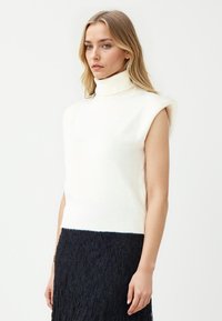 Ärmelloser weißer Rollkragenpullover aus weichem Strickmaterial, mit einer taillierten Passform und geripptem Saum, kombiniert mit einem strukturierten schwarzen Spitzenrock.
