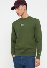 Mladý muž oblečený v zelenej mikine Tommy Hilfiger, modrých džínsoch a bordovom klobúku, stojí s jednou rukou v vrecku a pozerá sa doľava.