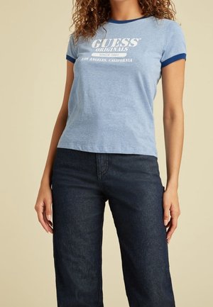 Kvinde iført en lyseblå Guess Originals T-shirt med mørkeblå kant og mørkeblå jeans, stående foran en beige baggrund.