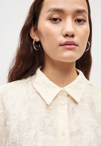 Kremowa koszula na guziki z teksturowanej, pogniecionej tkaniny, klasycznym kołnierzem i małymi, przezroczystymi guzikami. Wygląd dopełniają złote kolczyki-koła.