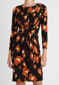 Robe noire à manches longues, avec des motifs abstraits orange et crème. Détail en nœud à la taille, silhouette près du corps et longueur au genou.