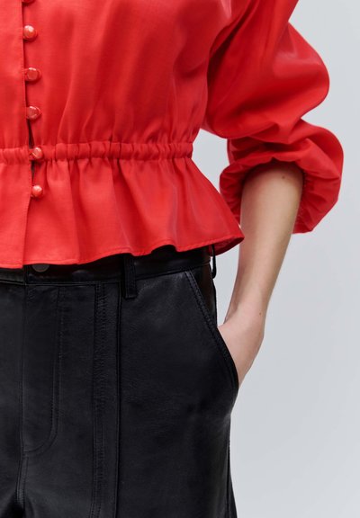 Kobieta w czerwonej bluzce z guzikami, marszczonym stanem i czarnych skórzanych spodniach, z ręką swobodnie wsuniętą do kieszeni.