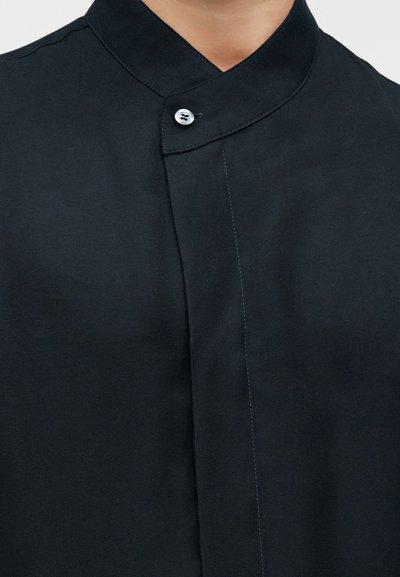 Κοντινό πλάνο μαύρου πουκαμίσου με γιακά mandarin που κλείνει με ένα μικρό λευκό κουμπί.